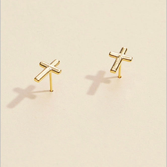 A pair of small gold cross stud earrings on a warm cream background with soft shadows.