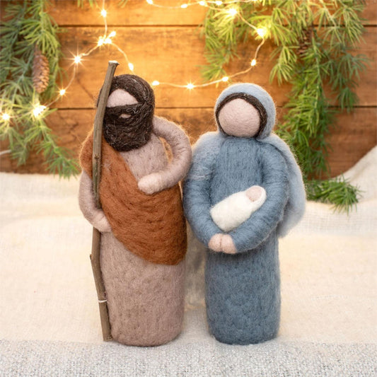 Two felted figures - Mary holding baby Jesus and Joseph.  Joseph in brown and Mary in blue, standing against a wooden background with lights and greenery.