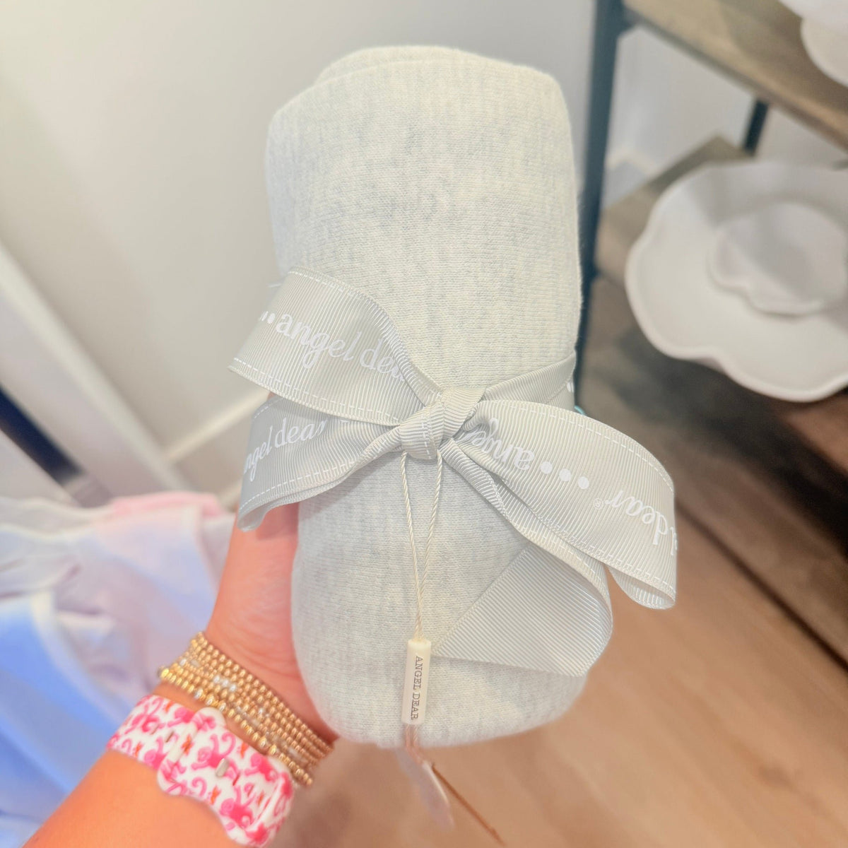 A light gray baby blanket rolled up and tied with a matching gray ribbon printed with the brand name 'angel dear', held by a hand with gold and pink bracelets.
