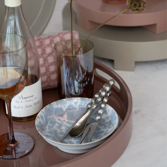 A styled tabletop scene with a round dusty-rose tray holding a blue marbled bowl, silver spoon and fork with beaded handles, a dark amber glass, and part of a wine bottle on a light marble surface.
