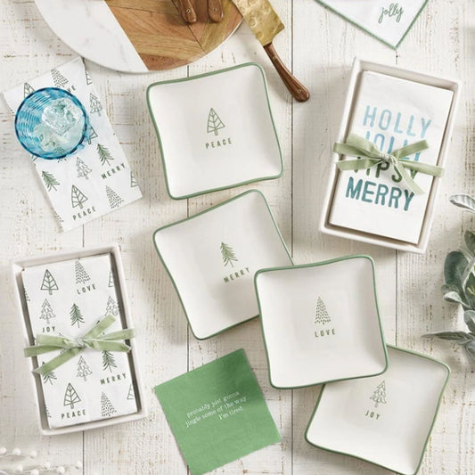 A styled flat-lay of white ceramic holiday-themed dishes and matching napkins with green tree prints and text reading Peace, Merry, Love, and Joy, arranged on a whitewashed wood surface with a blue glass, green ribbon, and small green card.