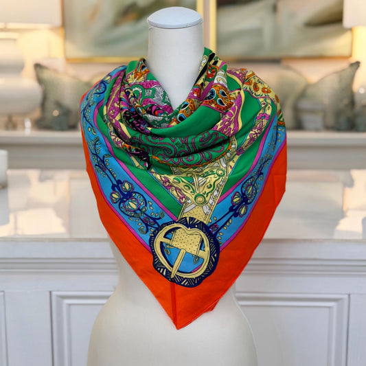 A silky square scarf in a bold equestrian print, styled around the neck with green, cobalt, magenta, and marigold patterns and an orange border.