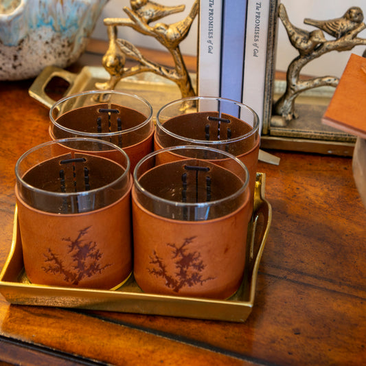 Four clear glass tumblers with brown sleeves and a tree motif, arranged on a gold tray on a wooden table, with decorative books and bird figurines in the background.