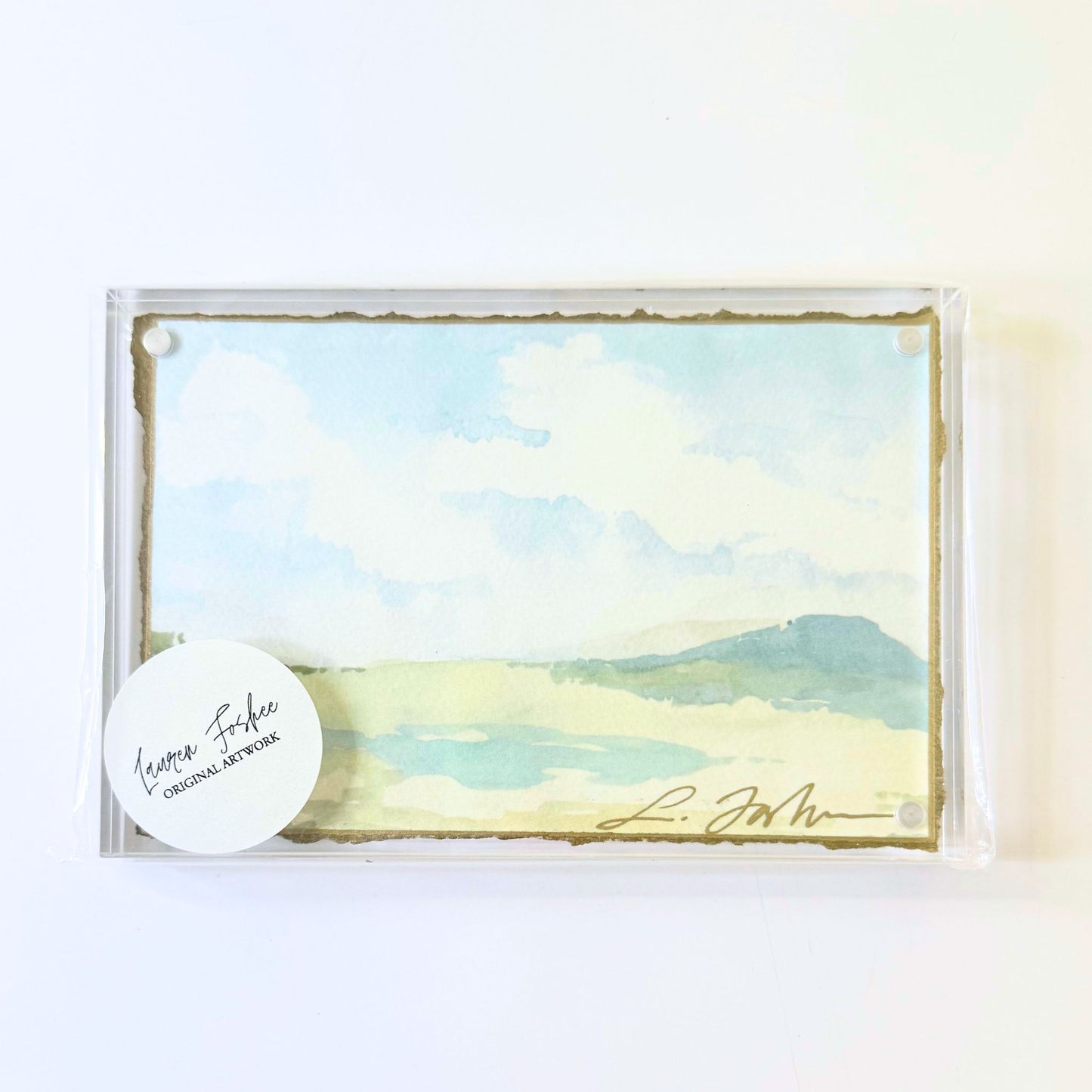 Lauren Foshee signed watercolor print, sky and clouds over a soft blue mountain and pale marsh, in a clear acrylic floating frame
