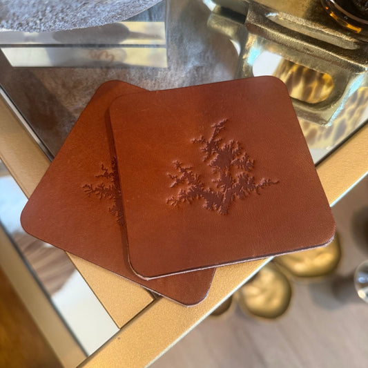 Two square brown leather coasters with rounded corners, stacked on a glass table. The top coaster features a stamp of Lake Martin, Alabama outline in a darker brown tone.