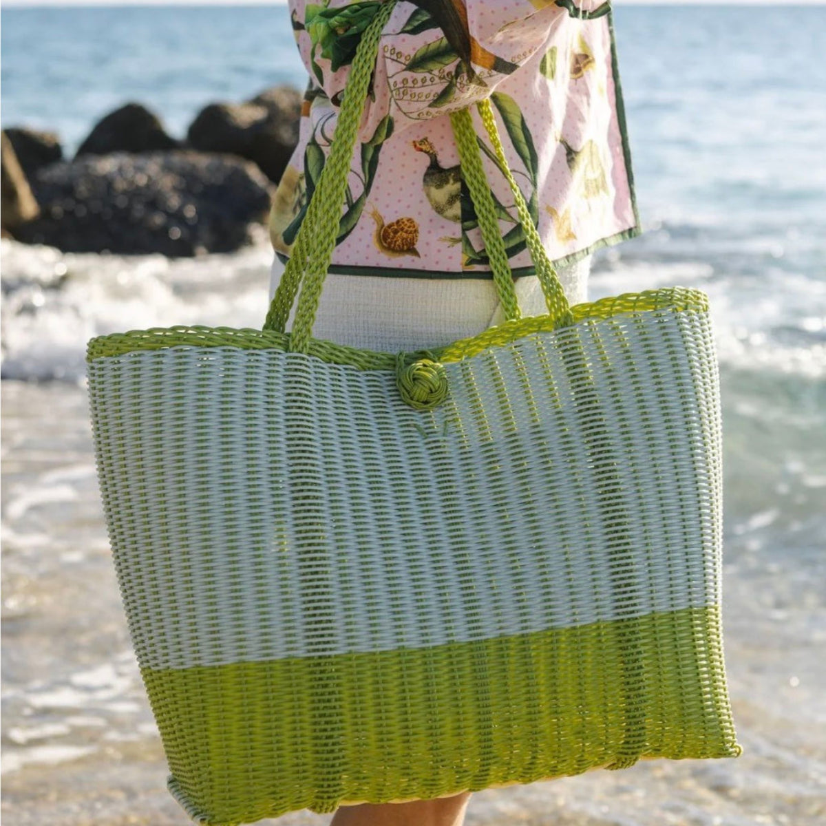 A large woven coated-plastic tote bag in white and lime green, carried on a beach by a person wearing a pink tropical print shirt.