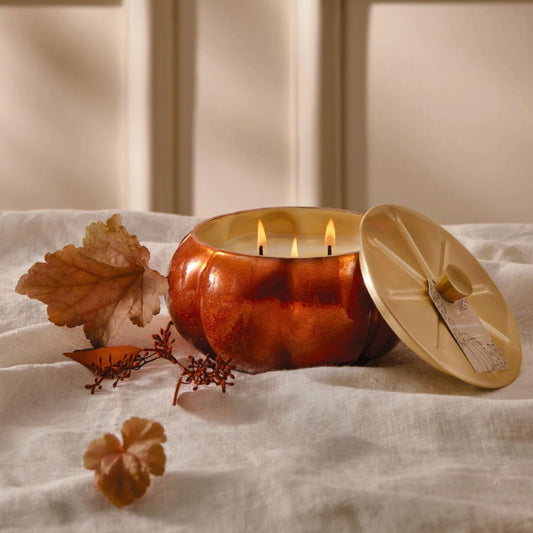 A lit three-wick pumpkin candle in a glossy amber glass jar with its matching lid leaning beside it on draped cream fabric, styled with autumn leaves and small sprigs in front of a softly blurred paneled background.