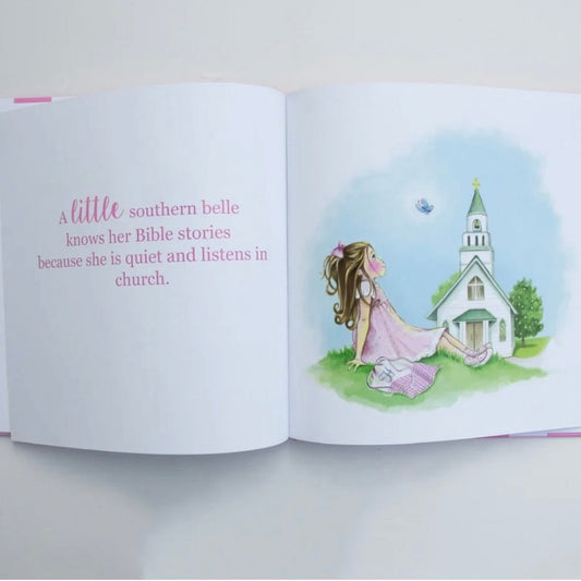An open children's book lying flat on a light gray surface. The left page has pink text reading, 'A little southern belle knows her Bible stories because she is quiet and listens in church.