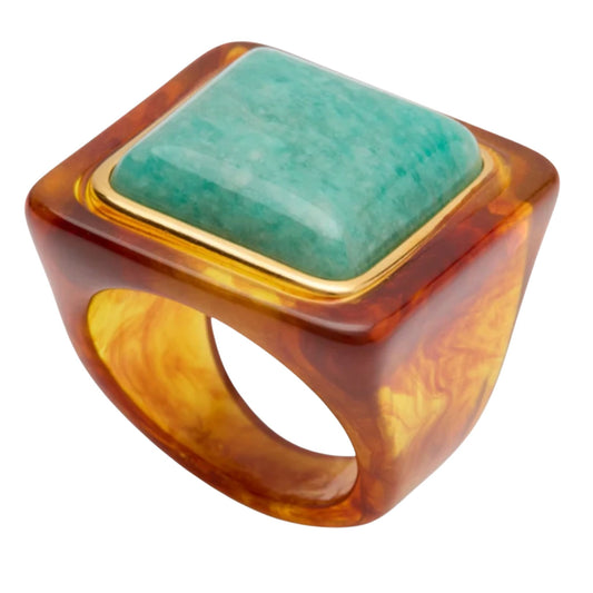 A square bezel ring featuring a smooth, polished light green stone inlay, surrounded by a gold-toned metal border and set within a thick, marbled amber-brown resin band.