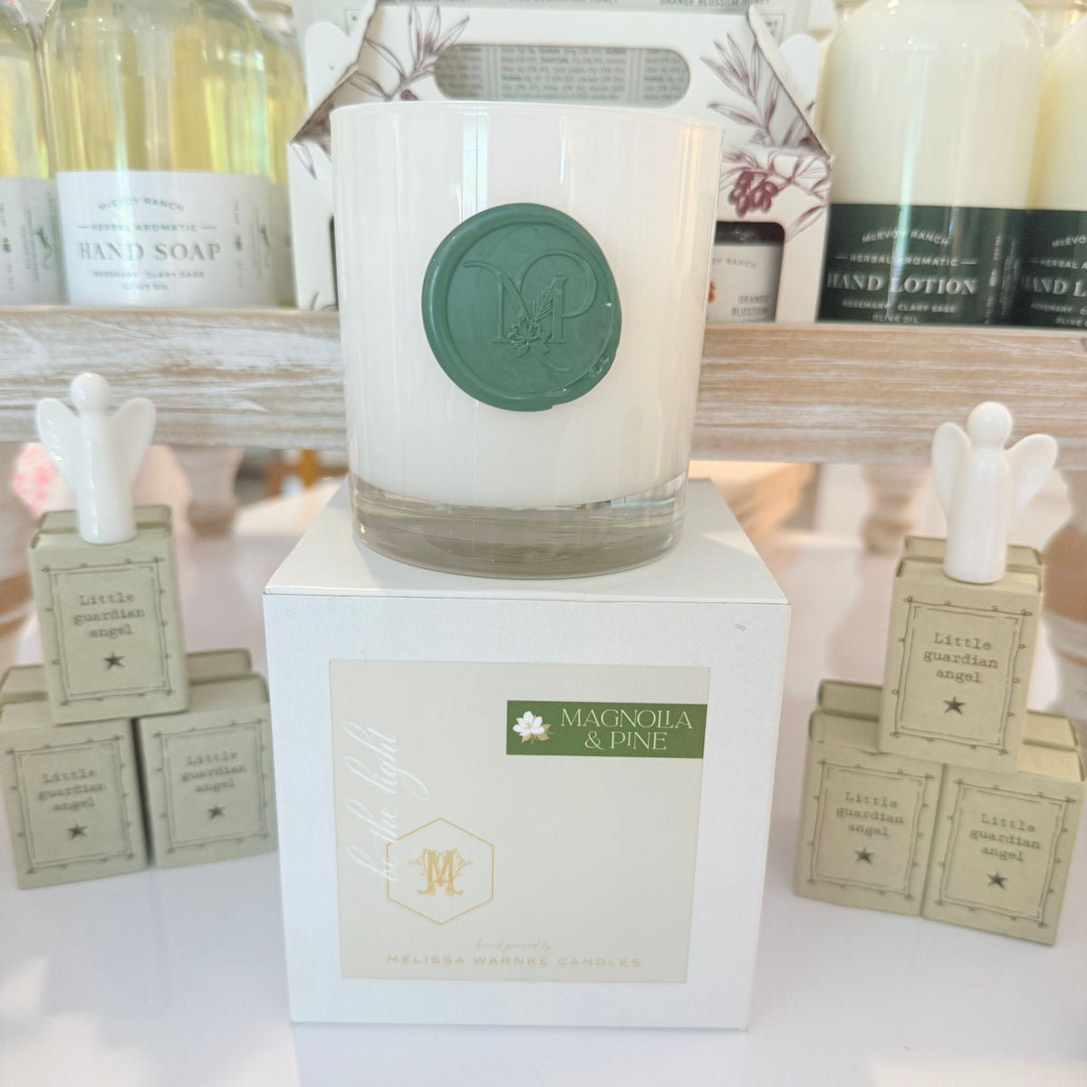 A white glass jar candle with a green circular wax seal sits on top of a white box labeled Magnolia & Pine. The candle is displayed in a styled retail setting with other bath and body products in the background.