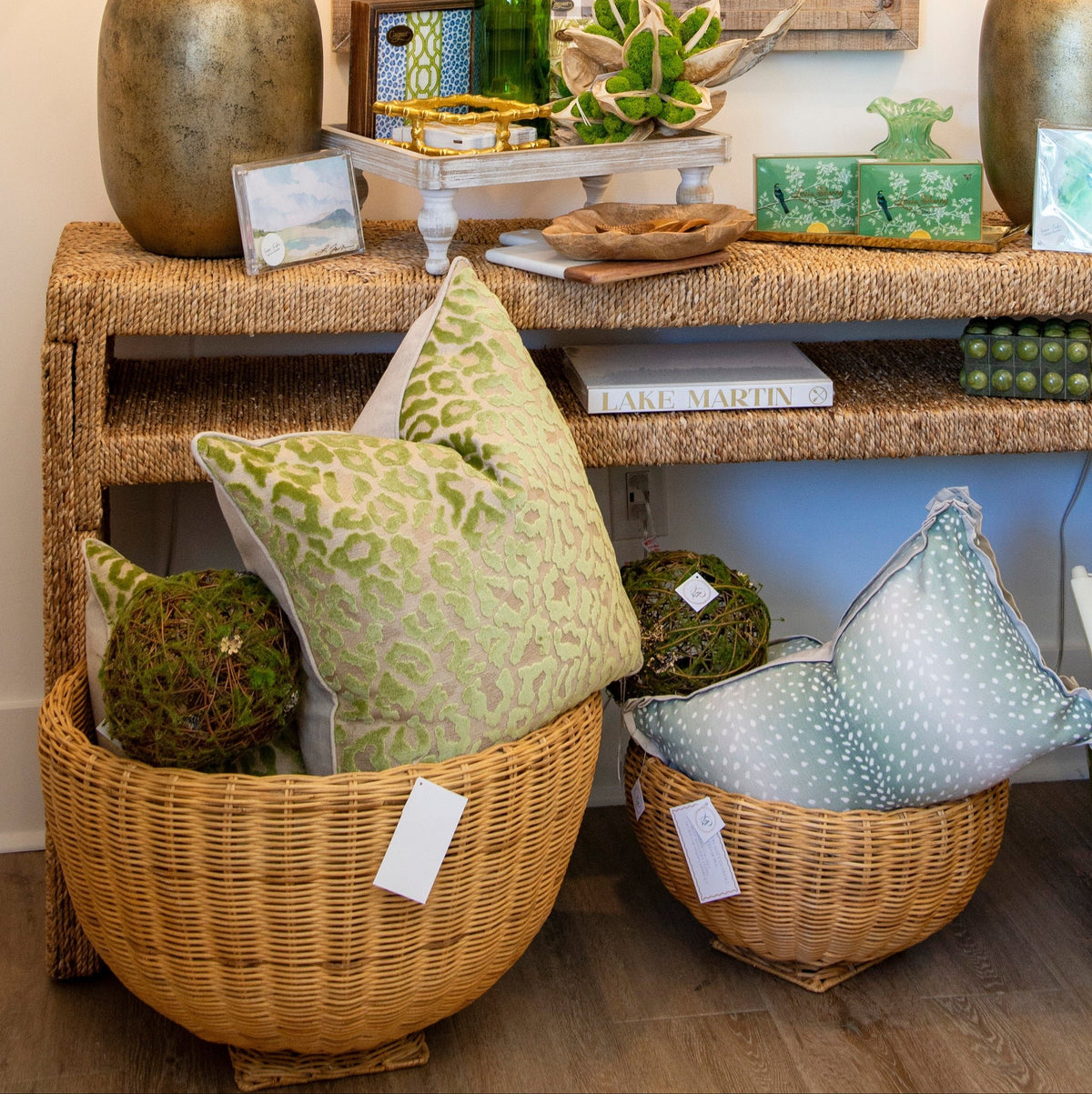 A styled home decor display featuring a rustic wood-framed mirror above a woven console table with two matching table lamps, green and blue patterned throw pillows in wicker baskets, and decorative green ceramic vessels on a wood floor.