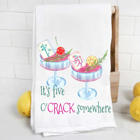 A white kitchen towel with a colorful watercolor print of two cocktails and the phrase 'It's five O'CRACK somewhere' in green and pink lettering. The towel is displayed hanging on a wooden rack against a white subway tile wall, with lemons on either side.