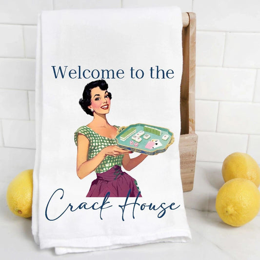 A white kitchen towel with a vintage-style illustration of a woman holding a tray, with the text 'Welcome to the Crack House' printed on it. The towel is draped over a wooden stand, with lemons and a white subway tile backsplash in the background.