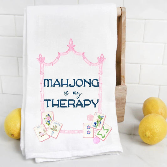 A white kitchen towel with a pink decorative frame and the text 'MAHJONG is my THERAPY' in navy blue, styled on a wooden stand with lemons against a white subway tile background.