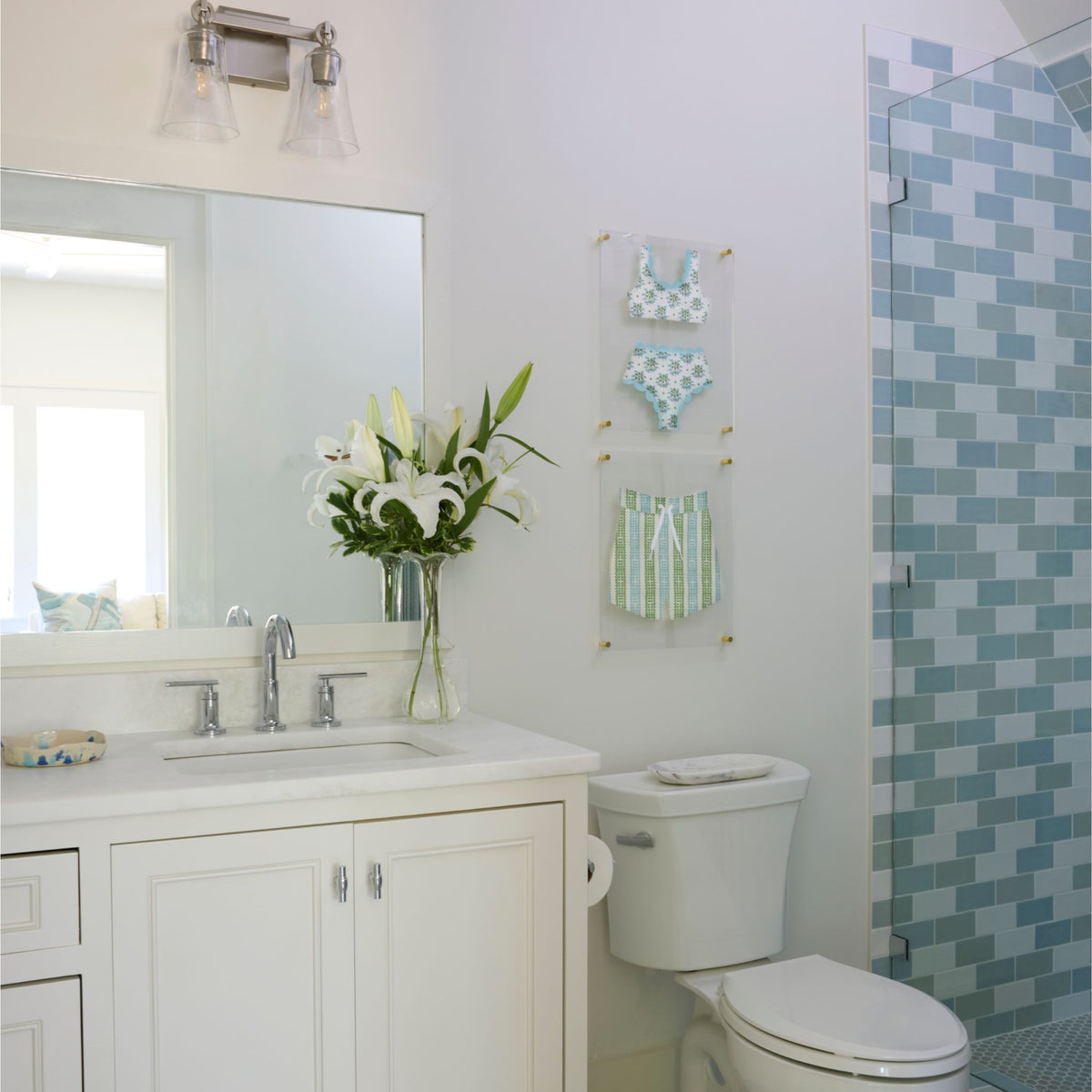 A bright, modern bathroom featuring a white vanity with a sink, a white toilet, a mirror, and a glass shower door with blue and green mosaic tile. Two clear acrylic wall frames hold fabric swimsuit art.  The top features a blue and white bikini, the bottom features a pair of blue, green and white swim trunks.
