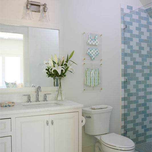 A bright, modern bathroom featuring a white vanity with a sink, a white toilet, a mirror, and a glass shower door with blue and green mosaic tile. Two clear acrylic wall frames hold fabric swimsuit art.  The top features a blue and white bikini, the bottom features a pair of blue, green and white swim trunks.