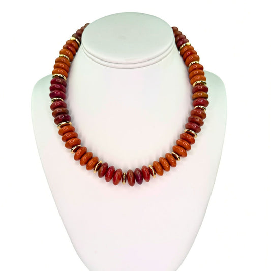 A beaded necklace made of polished oval beads in warm orange, red, and brown tones, separated by small gold spacer rings, displayed on a white jewelry bust against a white background.