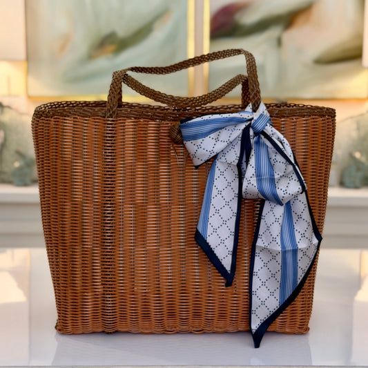 A brown woven wicker tote bag with two top handles, decorated with a blue and white patterned silk scarf tied in a bow on the front, set on a white shelf in front of a blurred background.