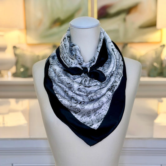 A black and white patterned square scarf with a solid black bow, displayed on a white mannequin bust in a softly lit indoor setting.