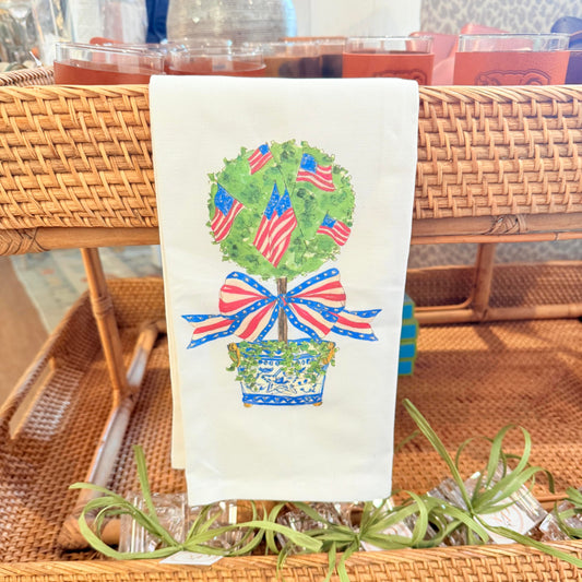 A white kitchen towel featuring a printed design of a potted topiary tree with American flags and a red, white, and blue bow, hanging on a wicker bar cart.