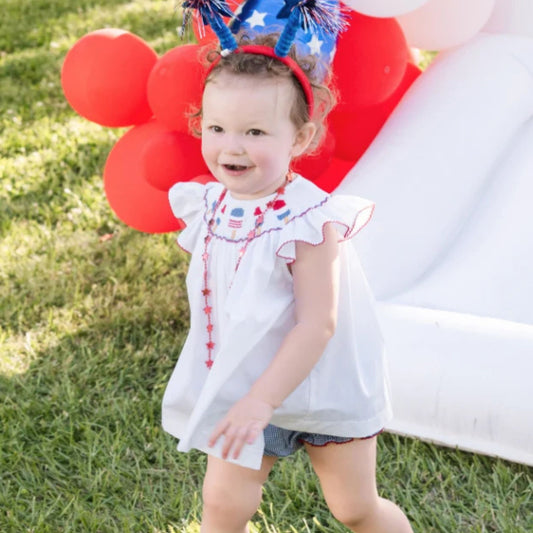 A smiling toddler in a white popsicle-smocked top and navy gingham bloomers stands on grass in front of red balloons at a Fourth of July party.