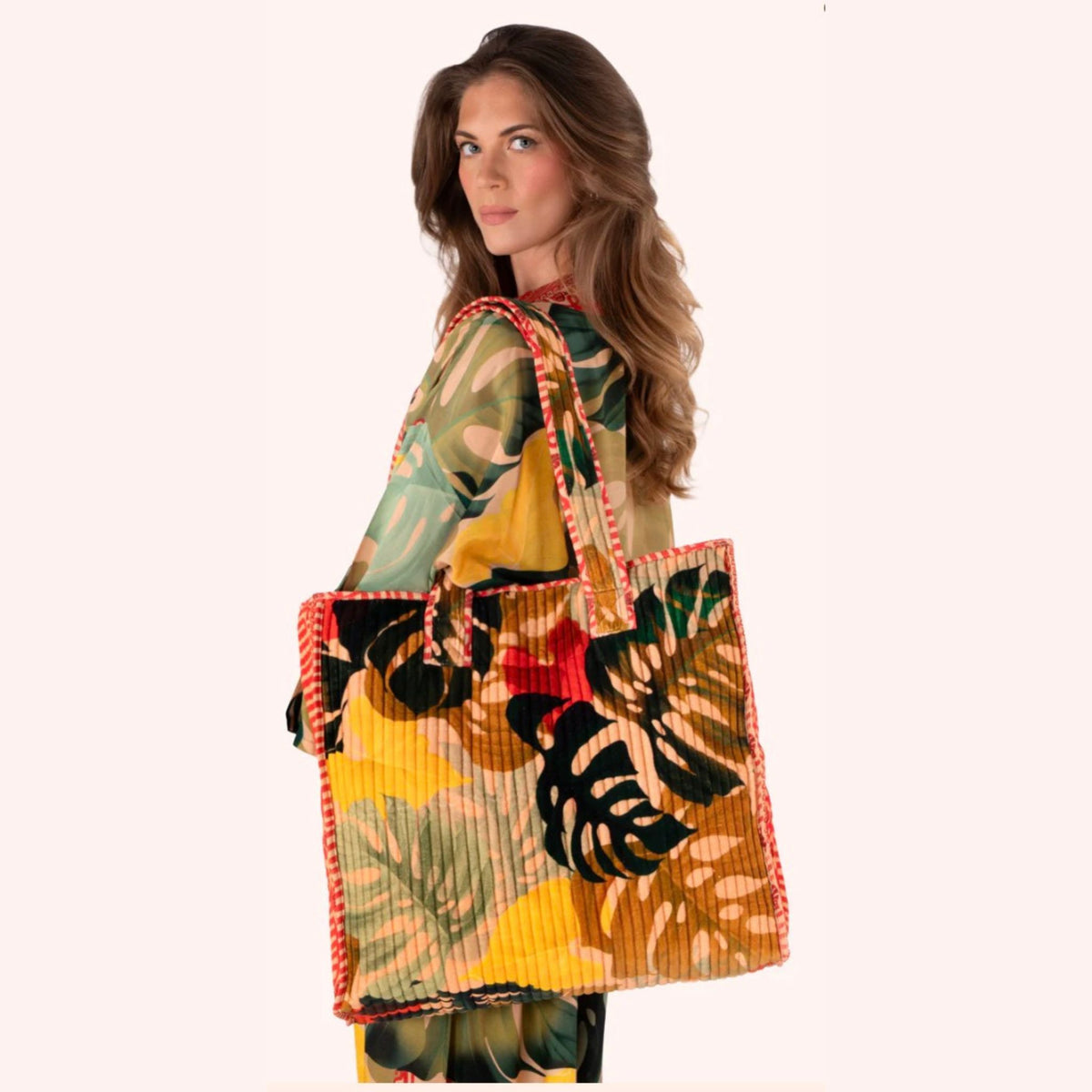 A woman wearing a tropical print top holds a large pleated tote bag with a matching tropical leaf print and red-orange braided handles, set against a solid pale pink background.