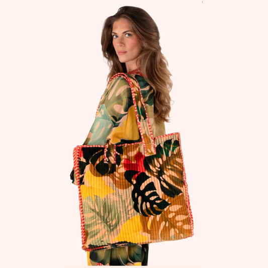 A woman wearing a tropical print top holds a large pleated tote bag with a matching tropical leaf print and red-orange braided handles, set against a solid pale pink background.