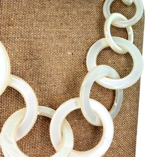 A close-up of a chunky link chain necklace made of milky translucent white shell or mother-of-pearl discs, draped over a brown burlap fabric background.