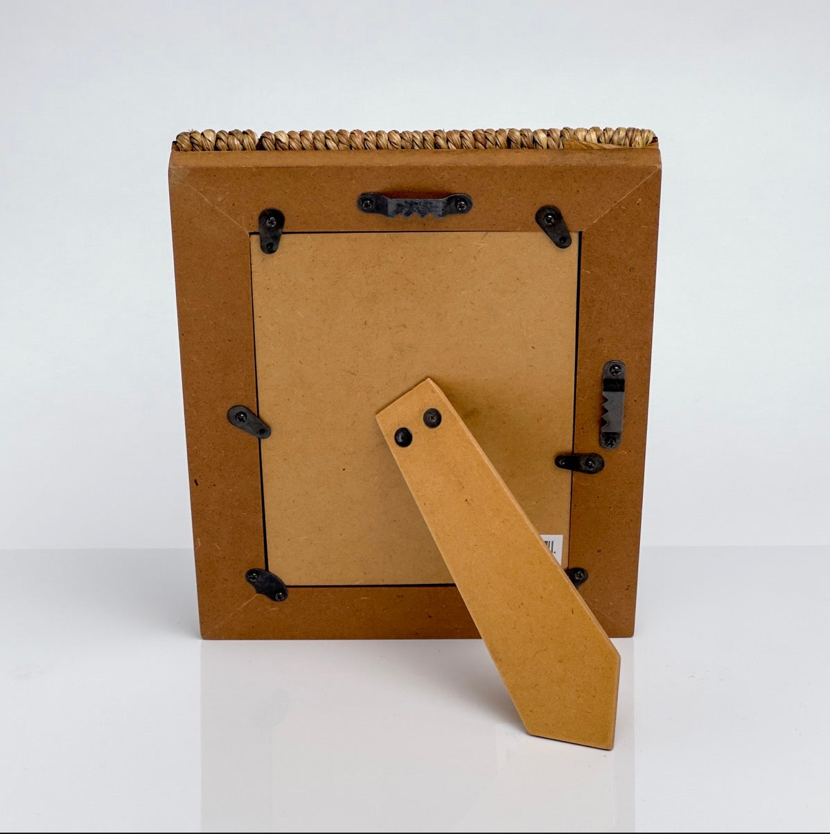 The back view of a square picture frame made from natural brown cardboard with a braided jute top border, secured with black metal clips and featuring a hinged cardboard stand.