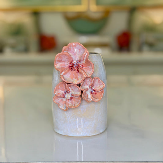 A cylindrical ceramic vase in a textured grey-white glaze, with three raised, sculpted flower blooms in blush pink on the front, set on a light marble surface.