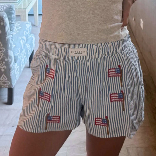 A person wearing Chakeris striped shorts with embroidered American flags, paired with a light gray t-shirt in an indoor setting.