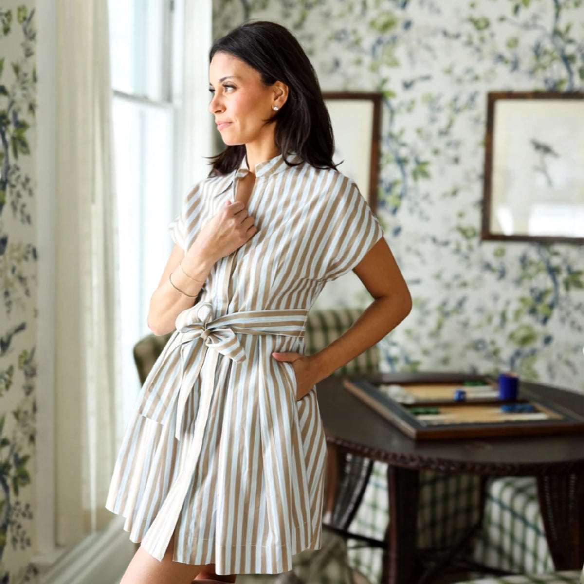 A woman wearing a short-sleeve tan striped shirt dress with a tie waist, standing in a room with floral wallpaper and a window.