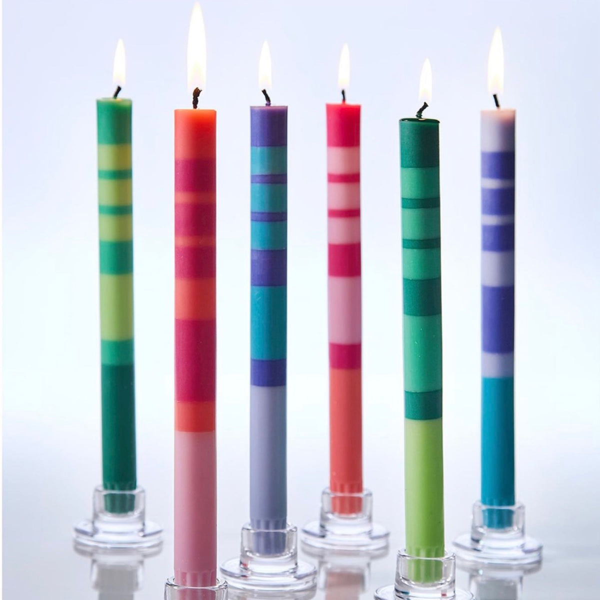 Six lit taper candles in stacked stripes of teal, red, purple, and green in clear glass holders