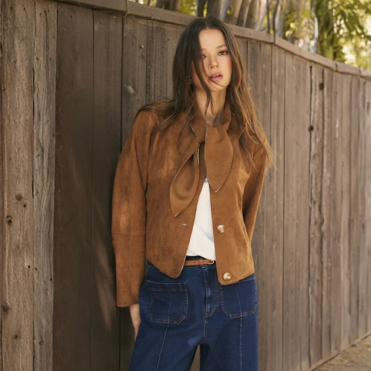 A model wearing a cropped camel faux suede jacket with a self-tie neckline, styled in a lifestyle setting.