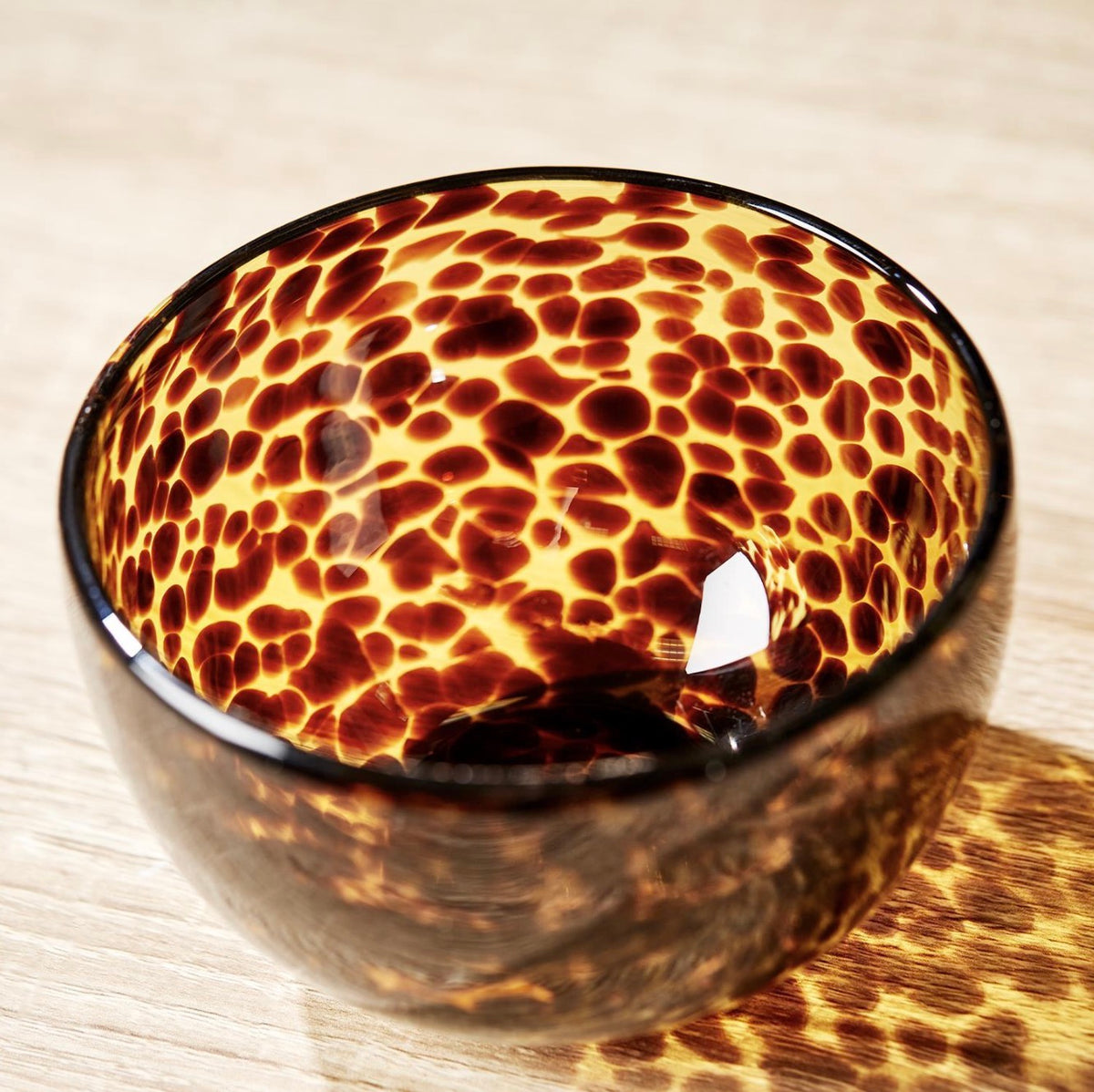 A close-up of a glossy bowl with a dark brown and amber mottled pattern, resting on a light wood surface.