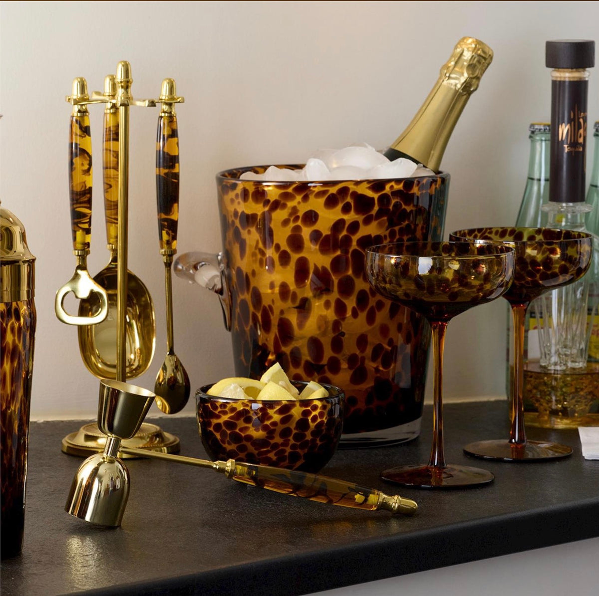 A bar-themed collection of amber and gold glassware and metal tools, including a leopard-print ice bucket with a champagne bottle, coupe glasses, a small bowl, and gold-toned bar accessories on a dark countertop.