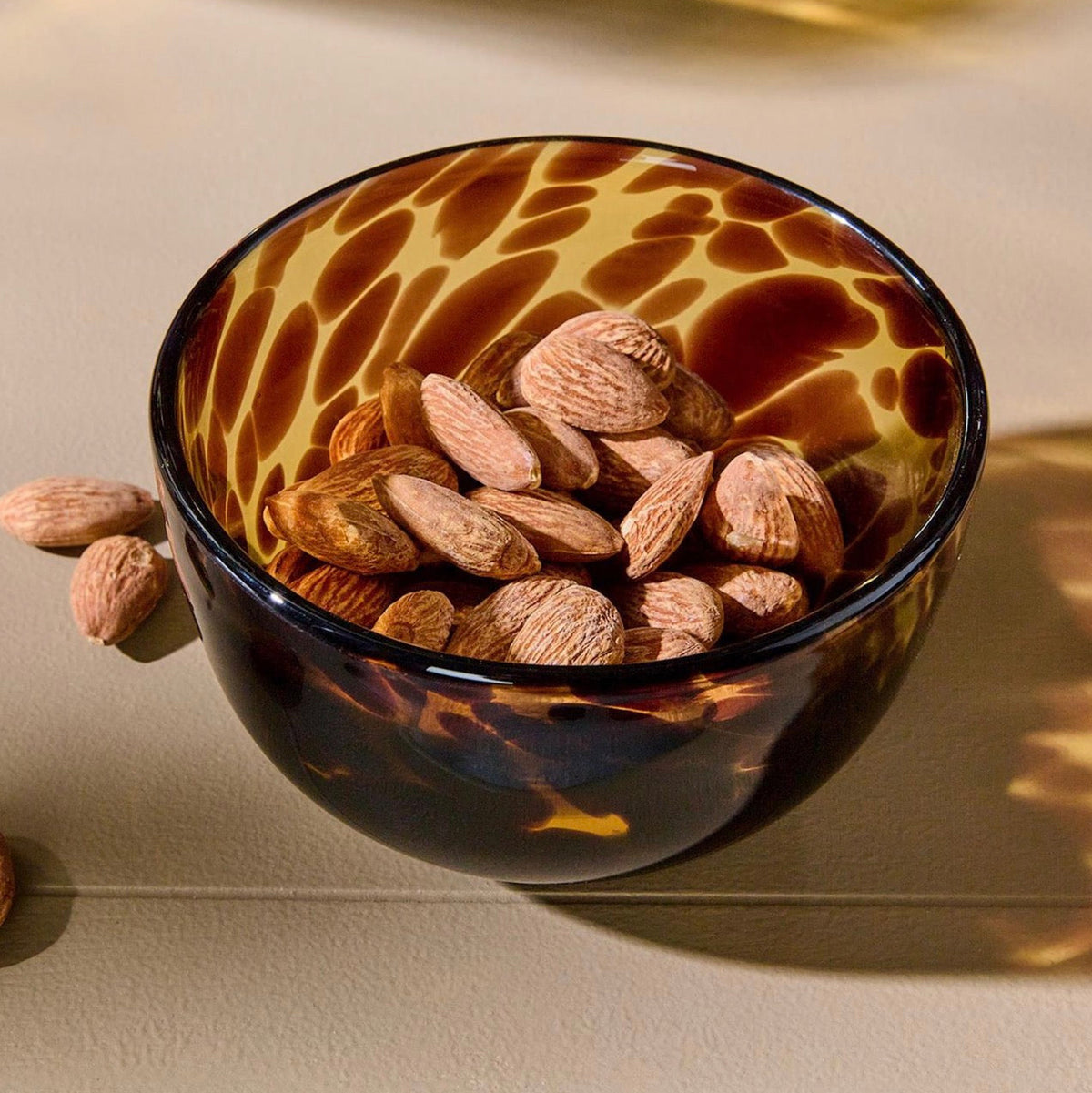 A small tortoiseshell glass bowl filled with almonds, photographed on a warm beige surface with soft shadows and light reflecting off the bowl.