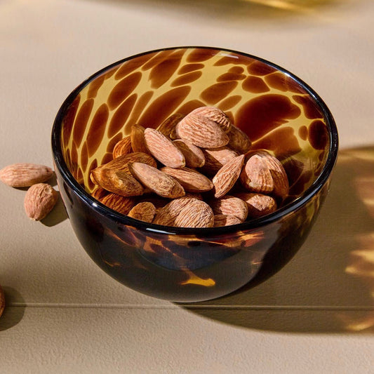 A small tortoiseshell glass bowl filled with almonds, photographed on a warm beige surface with soft shadows and light reflecting off the bowl.