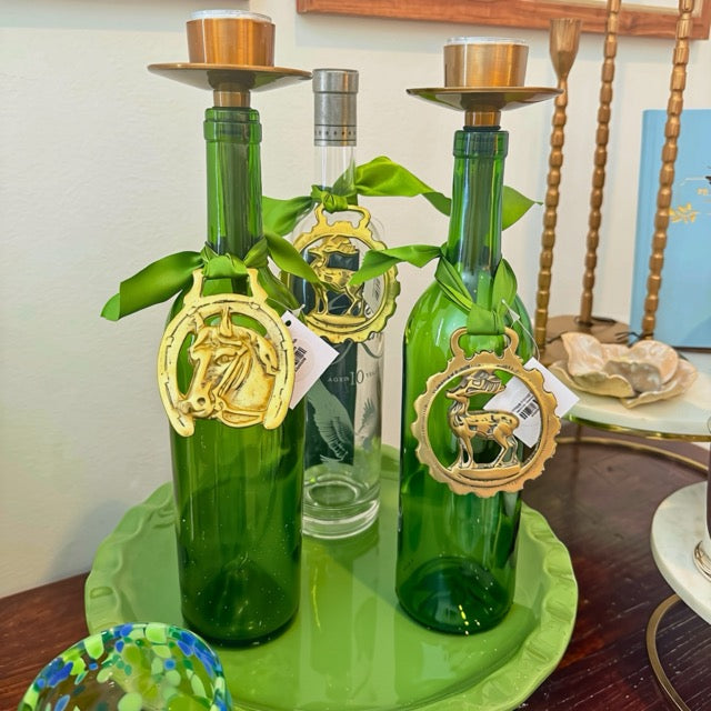 Two green glass bottle candlesticks with brass candle holders on top, decorated with green ribbons, gold horse and deer ornaments, and a small clear bottle in the center, displayed on a green tray.