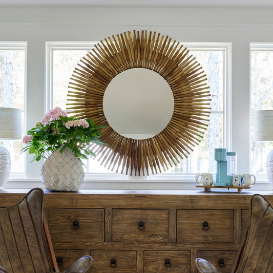 A large round wall mirror with a natural wood sunburst frame hangs on a white wall above a rustic wooden console table in a bright room. A white vase with pink flowers sits on the left, and mugs and a small appliance sit on the right.