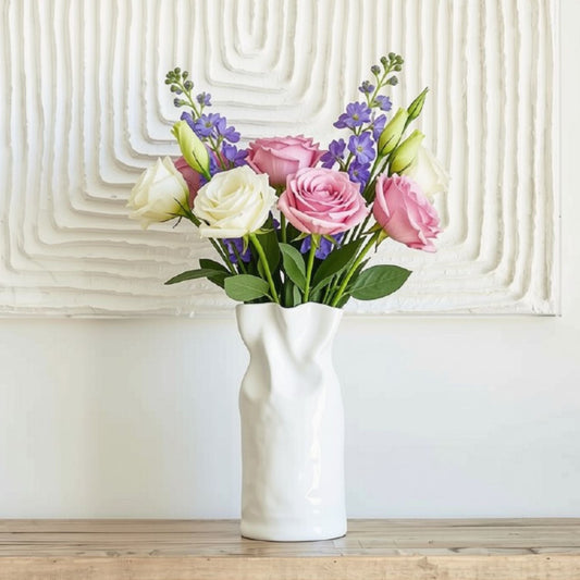A white ceramic vase holding a bouquet of pink and white roses with purple delphiniums and green leaves, placed on a light wood surface against a white wall with a textured geometric panel.