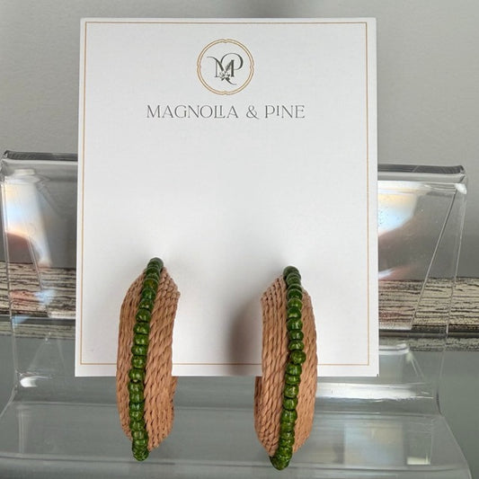 A pair of large hoop earrings displayed on a white Magnola & Pine card, set in a clear acrylic stand. The earrings feature a textured tan woven center with a border of small green beads.