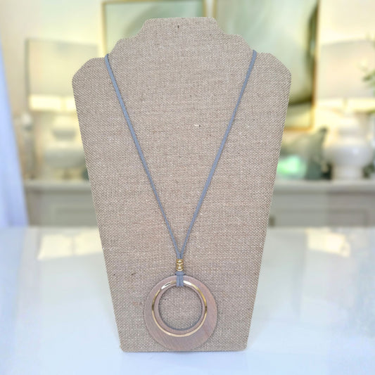 A circular pendant necklace displayed on a beige fabric-covered jewelry bust. The necklace has a muted blue-gray cord and a large circular pendant with a warm pinkish-gold metallic finish, set against a softly blurred indoor background.