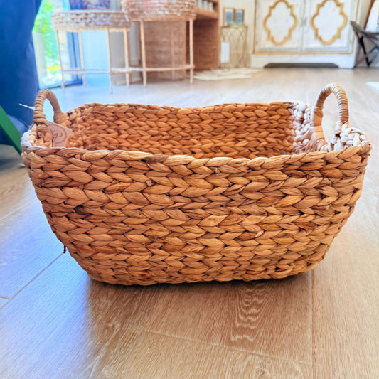 A rectangular woven basket made from natural brown seagrass or wicker, featuring two loop handles, placed on a light wood floor in a home interior.