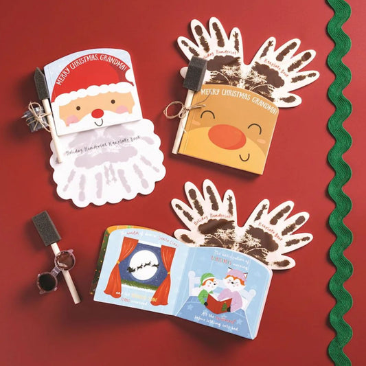 A flat lay of Santa and reindeer themed handprint keepsake books with foam brushes, paint, and an open children's book, on a red background.