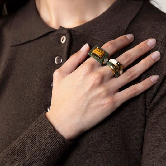 A hand wearing stacked resin rings — an ivory tiger eye ring, a jade and gold accent ring, and an ivory band — over a brown sweater.