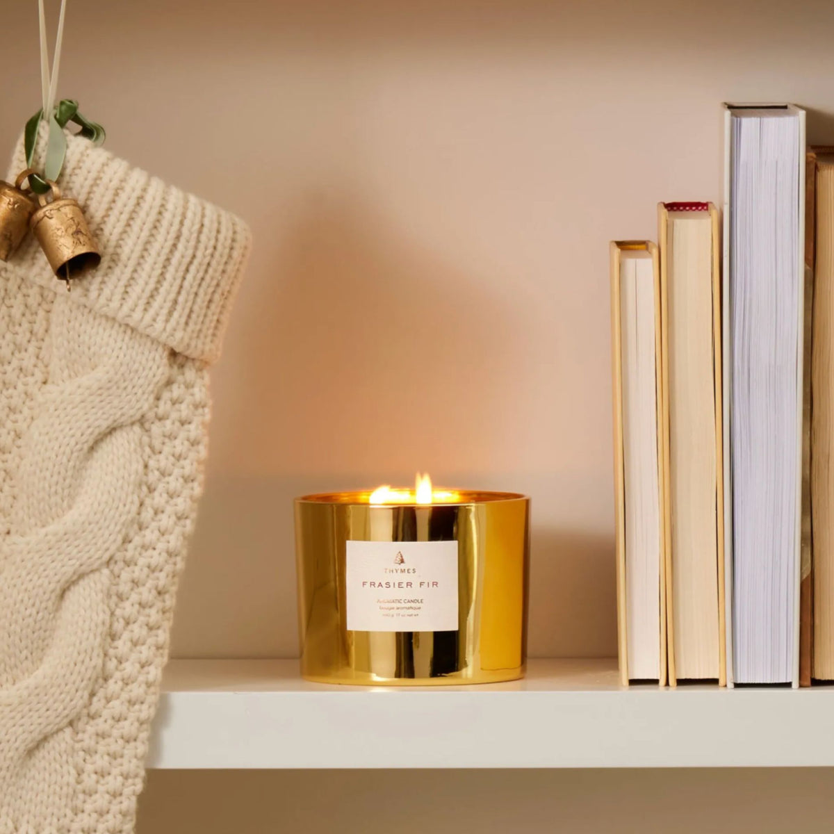 A lit gold Frasier Fir candle glowing on a shelf beside a cream knit stocking with bell ornaments and a stack of books.