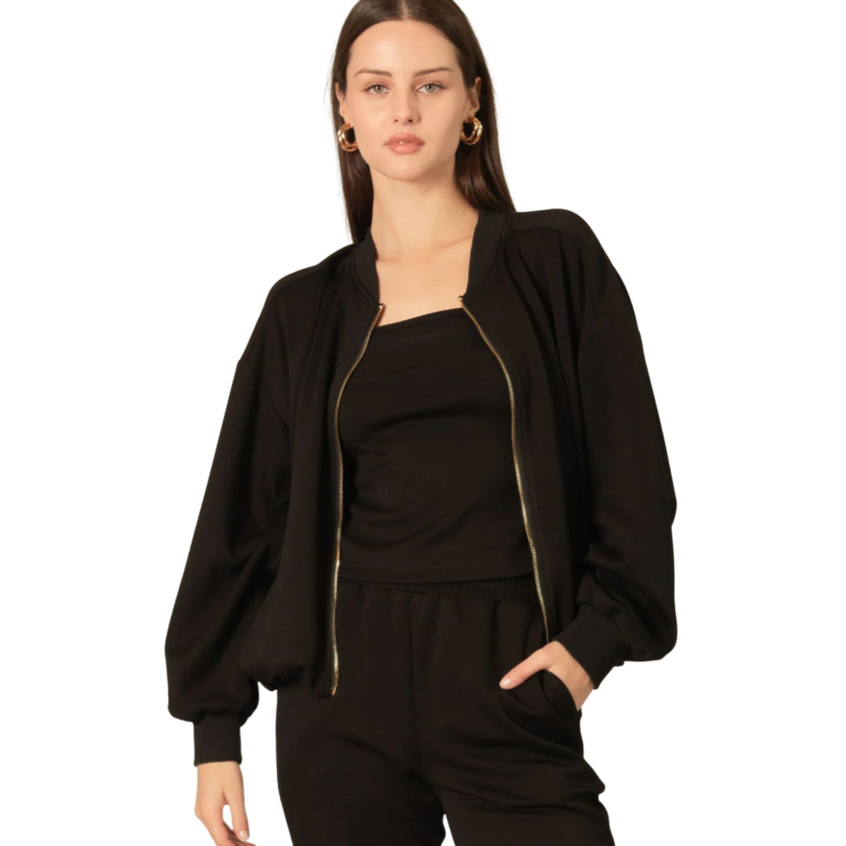 A woman wearing a black bomber jacket open over a black cami and black pants, with gold hoop earrings.