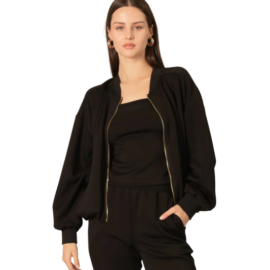 A woman wearing a black bomber jacket open over a black cami and black pants, with gold hoop earrings.