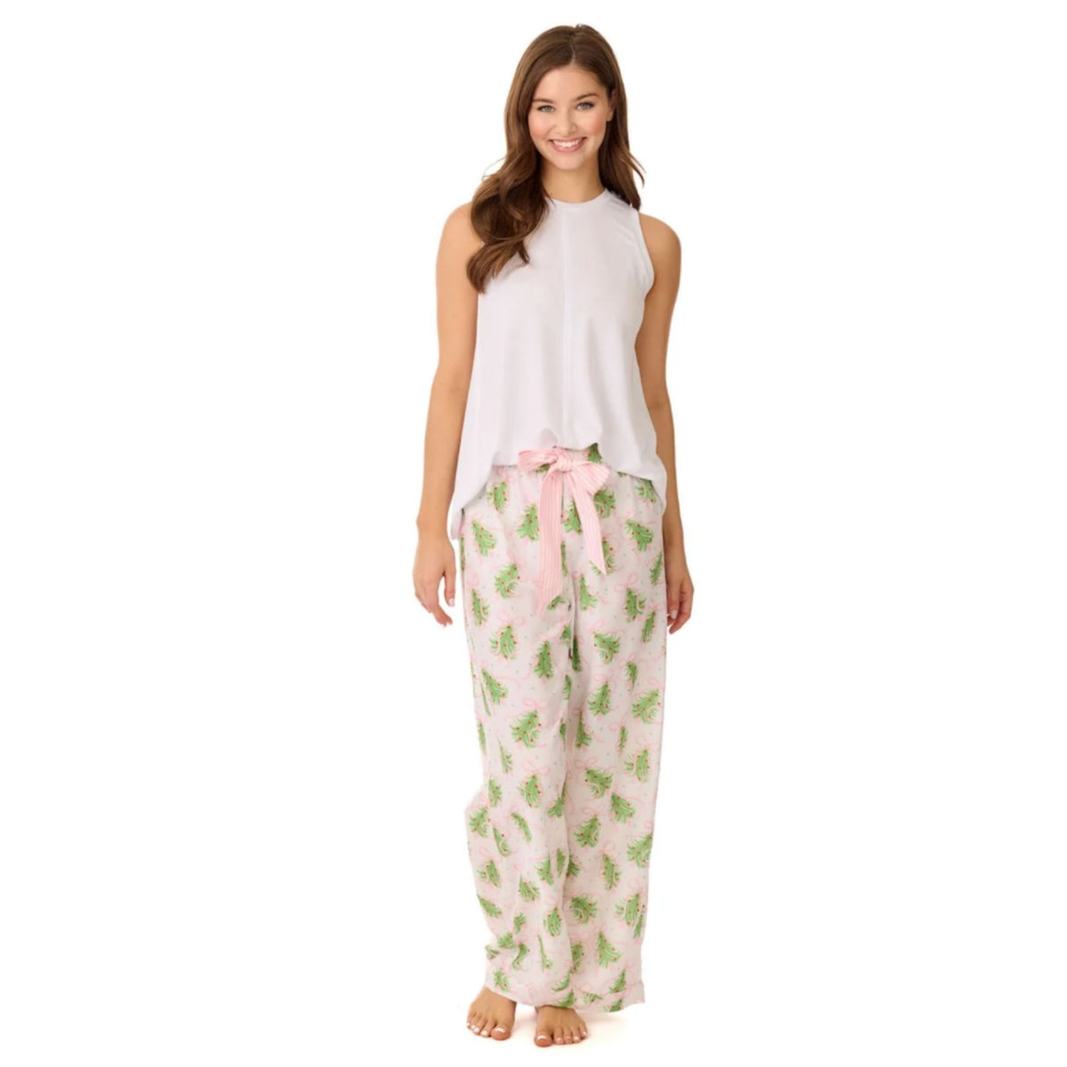 A woman wearing ivory pajama pants printed with green Christmas trees and pink bows, paired with a white sleeveless top.